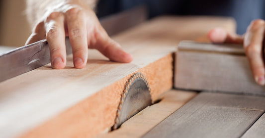 A toothed blade sticks up from a table and is used to cut through a light-colored plank of wood that's pushed along by hands.