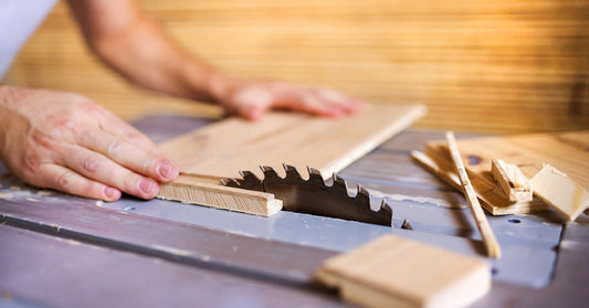 A handyperson lays a piece of plywood down onto a table saw and pushes the plywood through a saw blade.