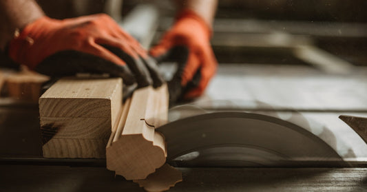 A carpenter uses a piece of wood to protect his hands as he uses it to push another piece of wood into a saw blade.