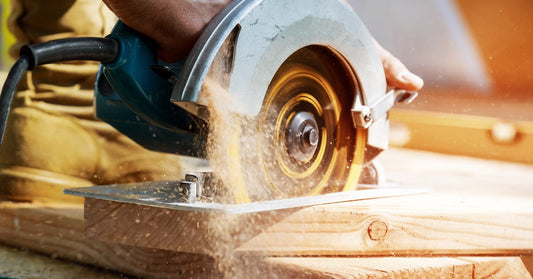 A pair of hands uses a circular saw to cut through a plank of wood, spraying sawdust behind it as it cuts.
