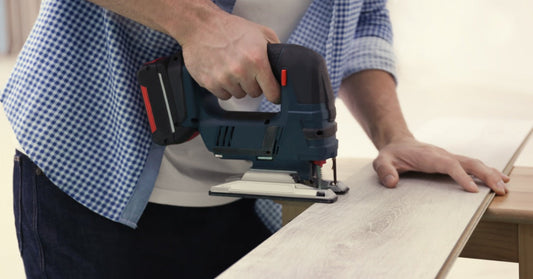 A man in a checkered shirt uses a jigsaw to cut through a board of laminate that's balanced on a table.