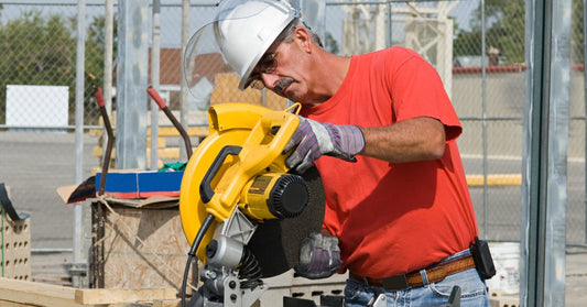 A construction worker in an orange shirt replacing the blade of a saw. There are several planks of wood next to him.