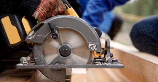 A builder in protective gloves using a handheld circular saw to cut through a piece of wood below them.