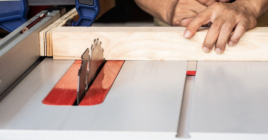 A carpenter's hands pushing a length of wood into a table saw. The saw blade is not moving and has a guard.