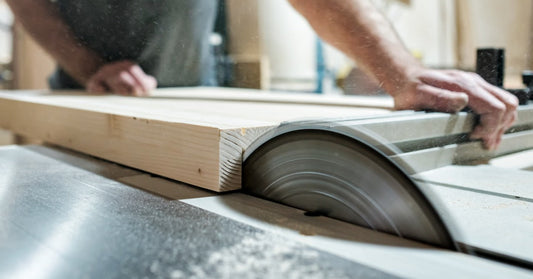 A man pushing a large piece of wood into a spinning table saw. The saw is cutting a channel into the wood.