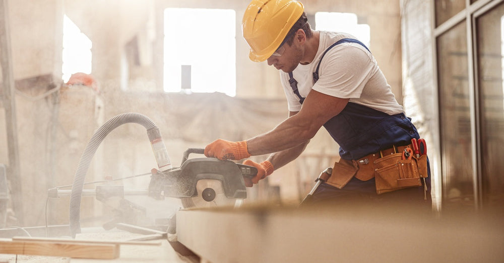 A construction worker wearing a hard hat using a circular saw to cut through a large plank of wood on his workbench.