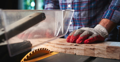 A person wearing thick gloves using a table saw by pushing a plank of wood close to the exposed blade.