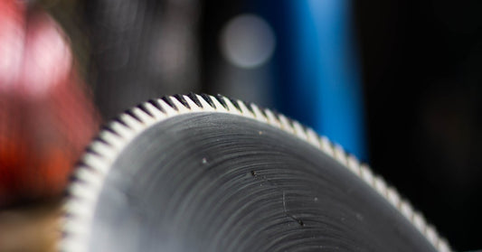 A circular saw blade with carbide tips glints, reflecting the light in front of a blurry background.